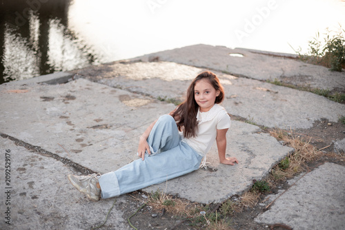 Fotografie A young girl of Hispanic descent sits casually on a sunlit dock, showcasing her relaxed style and playful spirit