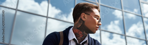Obraz na plátně A young man with tattoos and stylish glasses stands against a glass building reflecting clouds