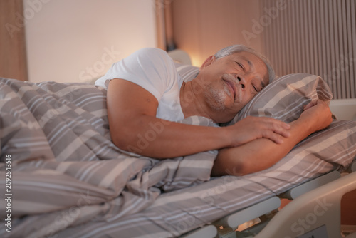 Nursing care: Elderly Asian man resting peacefully in a hospital bed as he recovers, eyes closed under soft warm light in a calm care room, a moment of gentle care