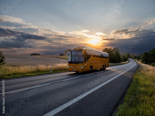 A yellow bus driving on the asphalt road winding through idyllic rural landscape during mystical sunset