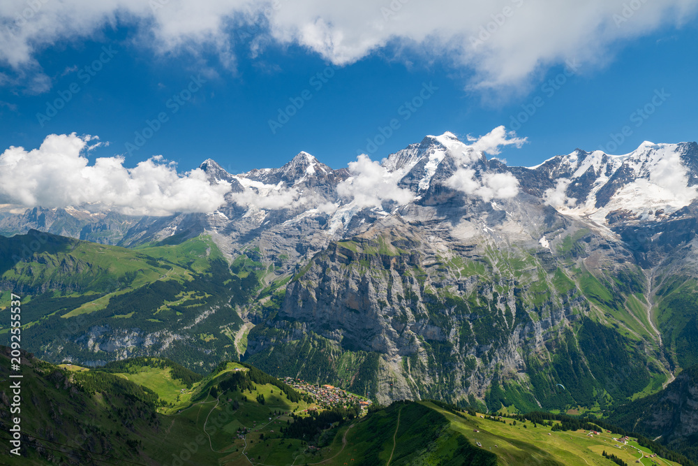 Fototapeta premium Wide view of Swiss Alps above Lauterbrunnen valley, with green slopes, rocky ridges, lingering snowfields, and summer clouds under blue sky.
