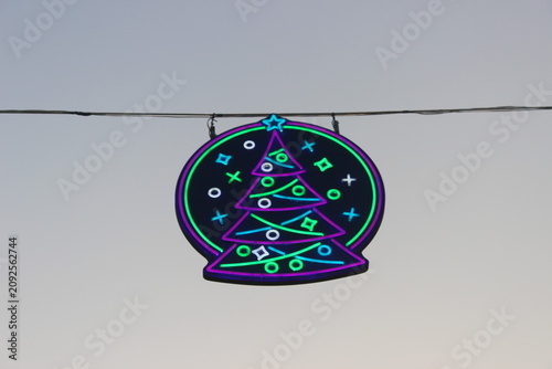 Neon christmas tree light display sign hanging against clear evening grey skies.