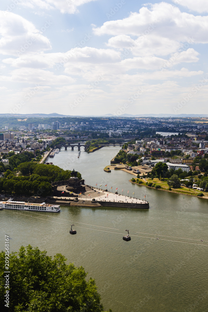 Obraz premium Elevated panoramic view of Koblenz with the Rhine and Moselle river confluence, Deutsches Eck, cable cars, bridges, and historic architecture beneath a dramatic summer sky.
