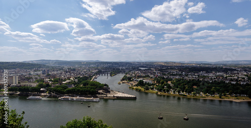 Photos Panoramic view of Koblenz with the Rhine and Moselle rivers, bridges, cable cars, and historic skyline beneath a bright summer sky with scattered clouds