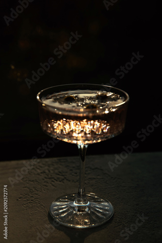 A crystal coupe glass filled with a cocktail catches bright golden light with dramatic light reflections on a dark background. Luxury drink, night party mood, mysterious atmosphere