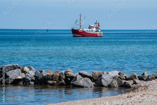 Fotografie Red Fishing Boat Navigating in Open Sea Seen From Sandy Beach at Baltic Sea