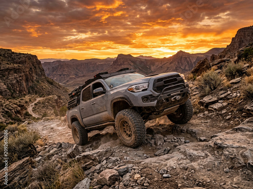 Grey pickup truck driving off road on rocky terrain during a bright sunset with orange sky and mountains in the background