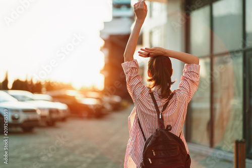 Young woman with a backpack walks away along a sidewalk at sunset, arms raised in joy, wearing a striped shirt and casual jeans as golden light washes the scene and creates a soft glow