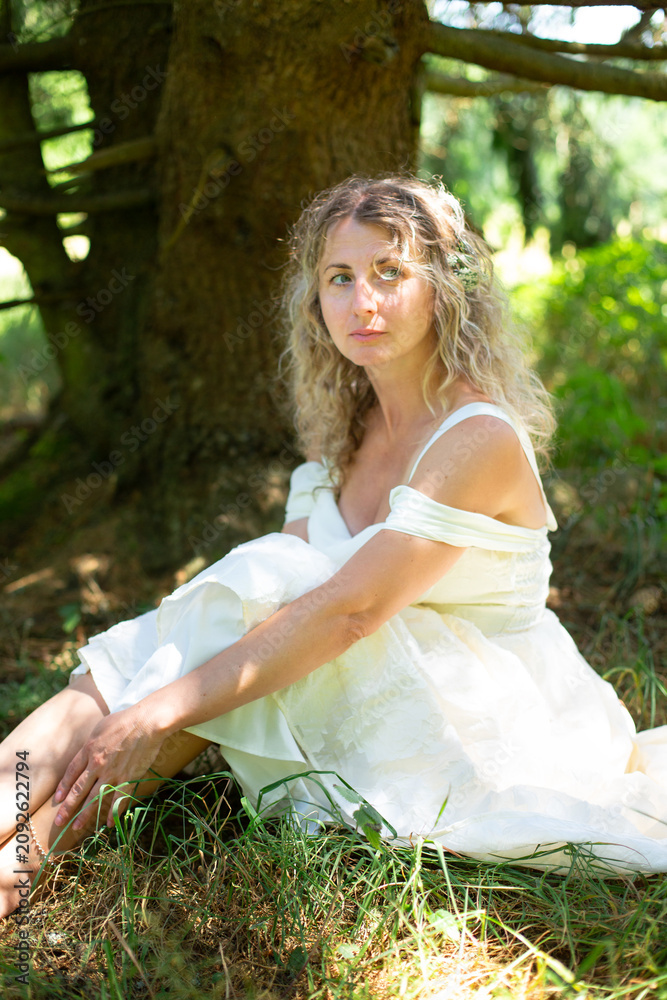 Fototapeta premium Blonde woman in a long white off-shoulder dress sits on the forest floor beneath the spreading branches of a large tree