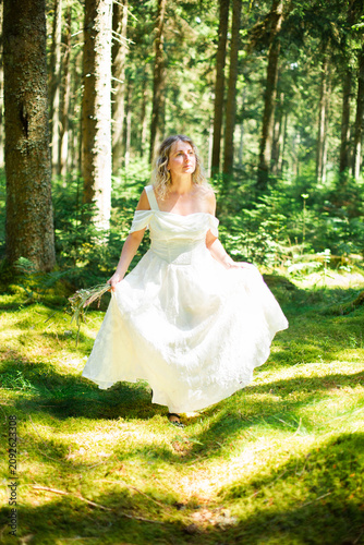 Fotografie Smiling blonde woman in a long white off-shoulder dress walks toward the camera while holding her flowing skirt in a sunlit green forest