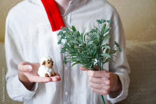 The young man's hands hold a miniature vintage figurine of a white sheep, the symbol of the year 2027 according to the Eastern calendar, against the background of a green bouquet of mistletoe 