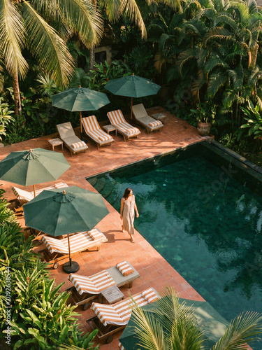 Woman walking beside a swimming pool with lounge chairs and umbrellas in a tropical setting
