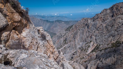 Vast and rugged landscapes of the Pamir mountains extend endlessly, showcasing steep gorges and jagged peaks under a clear blue sky. Adventurers roam the rocky terrain, exploring its beauty.
