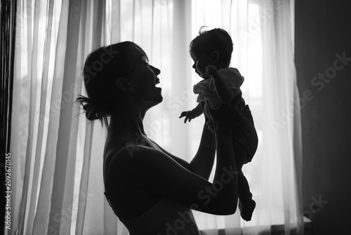 ilhouette of a mother lifting her baby up in front of a window, cozy home atmosphere. Black and white photo