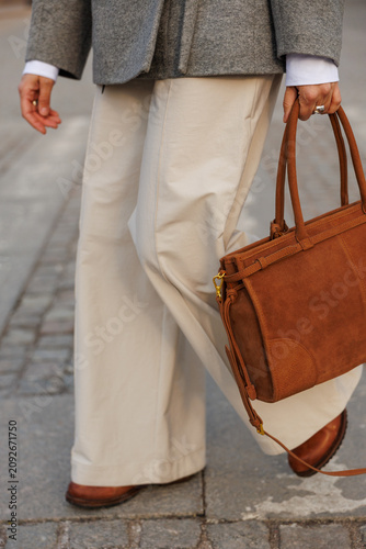 Woman striding across paved city road, carries brown suede bag in hand while walking. Grey wrap coat tied at waist over white trousers. Sunny daylight casts long shadow, relaxed urban street style.