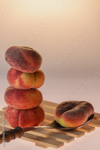 Flat peaches stacked on cutlery including fork and spoon on warm amber background. Playful food still life with kitchen props, studio composition, copy space.