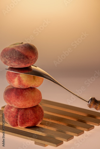 Flat peaches stacked in a tower with a knife resting alongside on warm amber background. Dramatic studio food still life with kitchen prop, copy space.