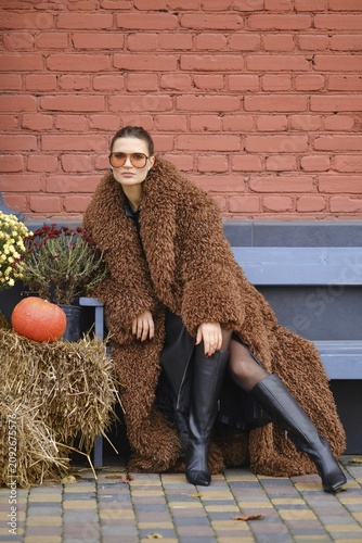 A woman wearing a brown fuzzy coat and knee-high black boots poses confidently outdoors. She is surrounded by autumn decorations, including a pumpkin, haystack and flowers, set against a brick wall
