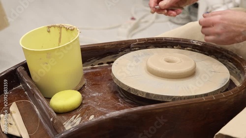 Close-up of pottery wheel with wet clay center, yellow water bucket, sponge, wooden trimming tools, and hands in background in a ceramic craft workshop studio for handmade artisan hobby.