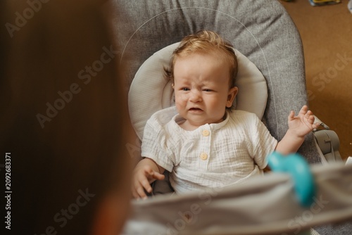 Photos Crying baby boy sitting in bouncer seat at home, upset toddler looking at mother