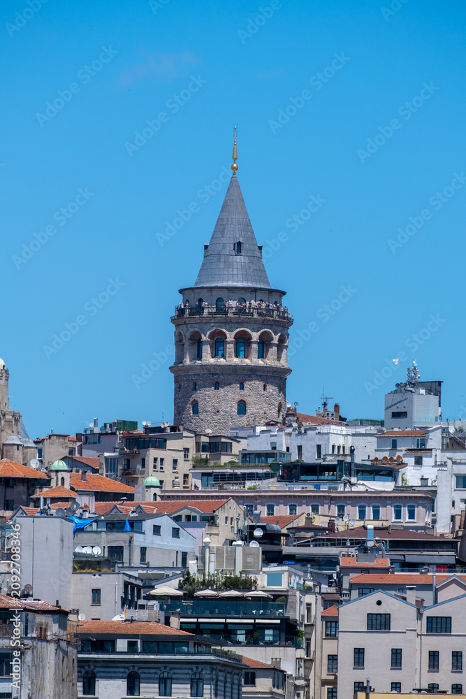 Obraz premium Galata Tower Rising Between Historic Buildings in Istanbul. Historic Galata Tower Surrounded by Istanbul Buildings. Galata Tower Emerging Above the Cityscape.