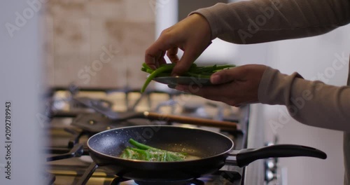 Hands are placing plate-held green beans flat into sizzling pan over blue gas flame to cook