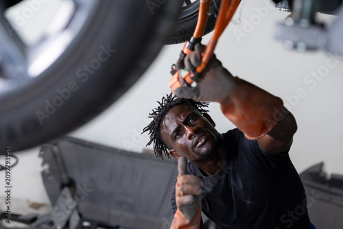 Fotografie Greasy auto mechanic working under a raised car, tightening parts with straps in a garage workshop, showing manual labor, focus, and automotive repair in progress