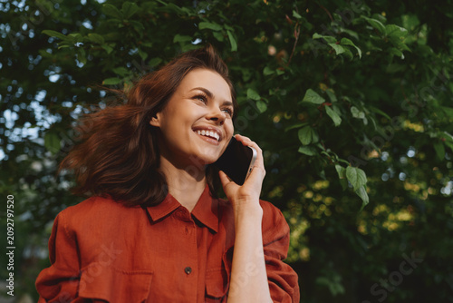 Cuadro en lienzo A smiling woman on a phone call outdoors in a bright orange jacket, framed by lush greenery
