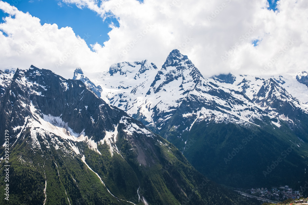 Obraz premium Majestic snow-capped mountains tower under a blue sky with clouds, revealing rugged terrain and green valleys in the foreground