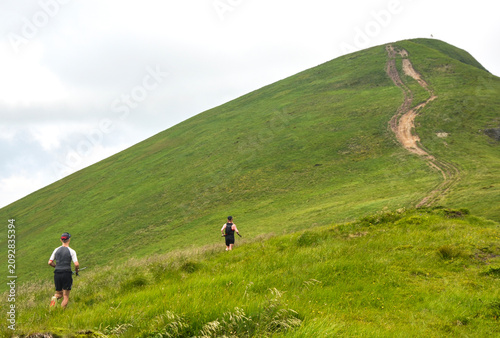 Athletes participate in a mountain trail marathon, running up a steep, vibrant green hill towards a peak under a cloudy sky. A dirt track winds along the ridge in a scenic alpine race