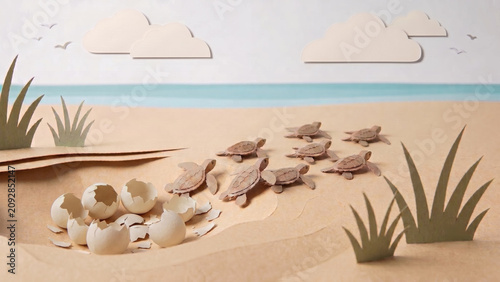 A construction paper diorama of baby sea turtles hatching from eggs and crawling across a sandy beach toward the ocean under a light sky with clouds.