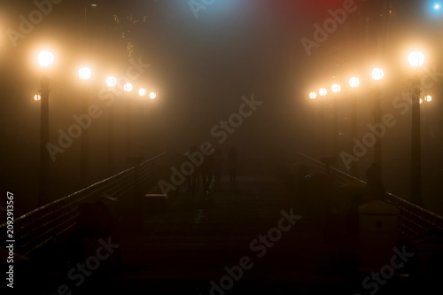 Silhouettes of people walking on a foggy pier with glowing streetlights at night