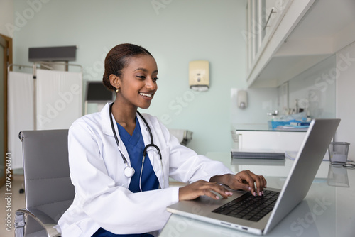 Tableau sur toile Happy young African general practitioner woman working at computer in medical of