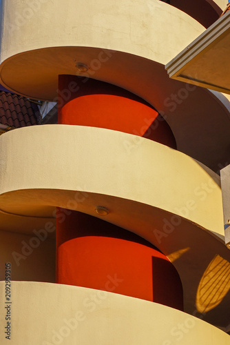 Modern spiral staircase with curved white concrete railings and bold red central columns, illuminated by warm golden evening light. Architectural details, geometric shapes, and dramatic shadows create