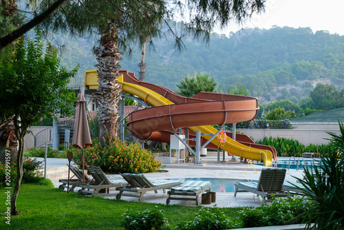 Outdoor swimming pool with colorful water slide, sun loungers and umbrellas at a modern resort hotel in Turkey. Summer vacation destination with mountains in the background and relaxing atmosphere