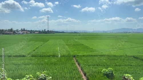 Beautiful countryside view from a moving train featuring lush green rice paddies, distant mountains, and a clear blue sky. A relaxing travel landscape in rural Indonesia.