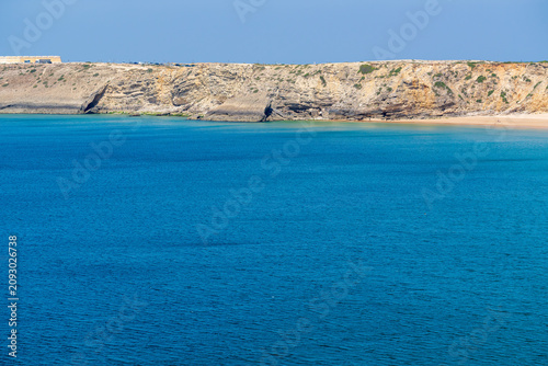 Vivid blue ocean waters stretch beneath dramatic layered limestone cliffs at a quiet bay along Portugal's Algarve coastline. Sunlight highlights the rugged rock and hidden sandy shore below