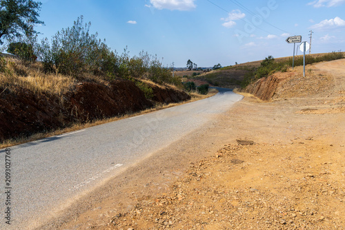 A quiet countryside road winds between dry grassy slopes and exposed reddish earth in Portugal's rural landscape. A dusty gravel junction with a road sign branches off under a warm summer sky
