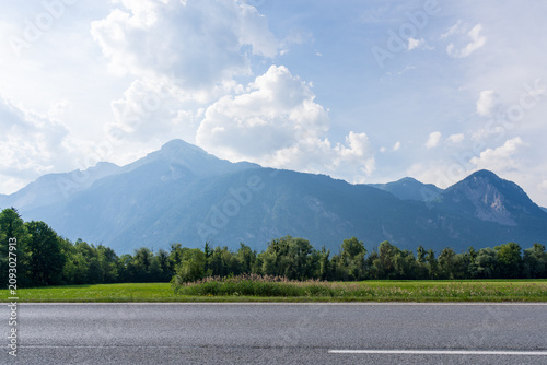 Hazy Alpine peaks rise above lush green meadows and blooming wildflowers in the Austrian countryside. A quiet rural road stretches beneath forested green hills under a cloud-filled summer sky
