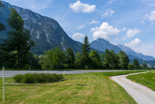 Towering Alpine peaks rise above a scenic highway and a winding paved bike path through a lush green valley in Austria. Fluffy white clouds drift over the mountains on a clear, sunny summer day