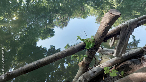 Tableau sur toile Rustic wooden branch fence by calm river with tree reflection