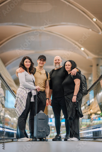 Group of four adults posing together on an escalator in a modern airport terminal, with bright lighting and a spacious interior visible in the background