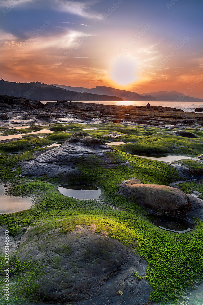 Obraz premium Golden sunset over Lastron beach in Bizkaia Spain with green mossy rocks during low tide, dramatic sky reflecting on the atlantic ocean coast