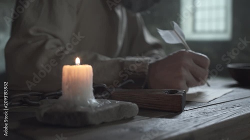 Close up of wooden inkwell on a rustic table against a blurred background of a medieval prisoner with chained hands writing with a quill pen inside a dark jail cell