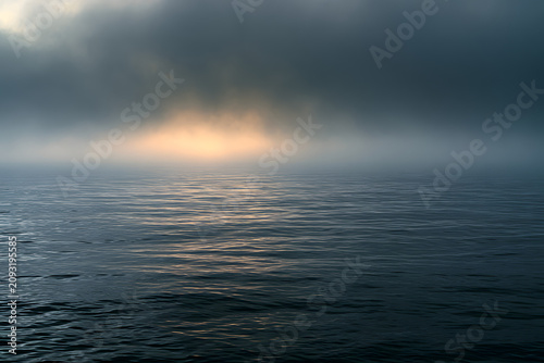 Golden light breaks through dark, misty clouds over a calm ocean, creating stunning reflections on the water surface. A serene yet powerful natural scene.