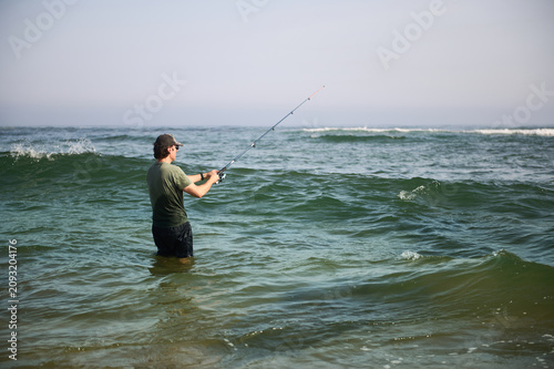 Foto Man fishing in the ocean waist-deep at the beach with a fishing rod on a calm da