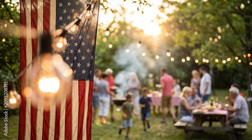 American flag waving softly in evening light with blurred backyard barbecue scene featuring family and friends eating and talking under string lights
