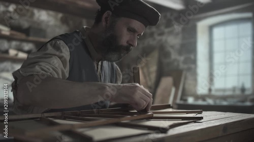 Medieval master testing the angles of an ornithopter wing frame with a wooden square on a rustic table inside a workshop