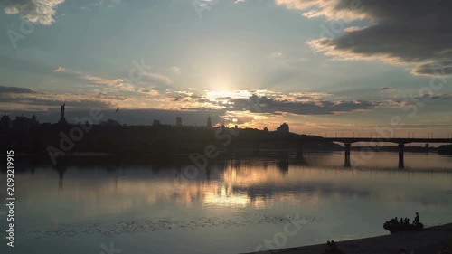 Sunset light breaks through clouds over a wide river Dnieper as a Kyiv city skyline and bridge form dark silhouettes, with calm reflections and a small boat drifting near the shore