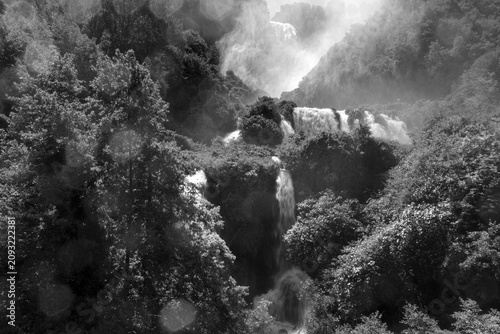 Vertical black and white view of Marmore Falls in Umbria, Italy. Multiple waterfalls cascade through dense forest while mist rises from the powerful water, creating a dramatic and timeless natural lan